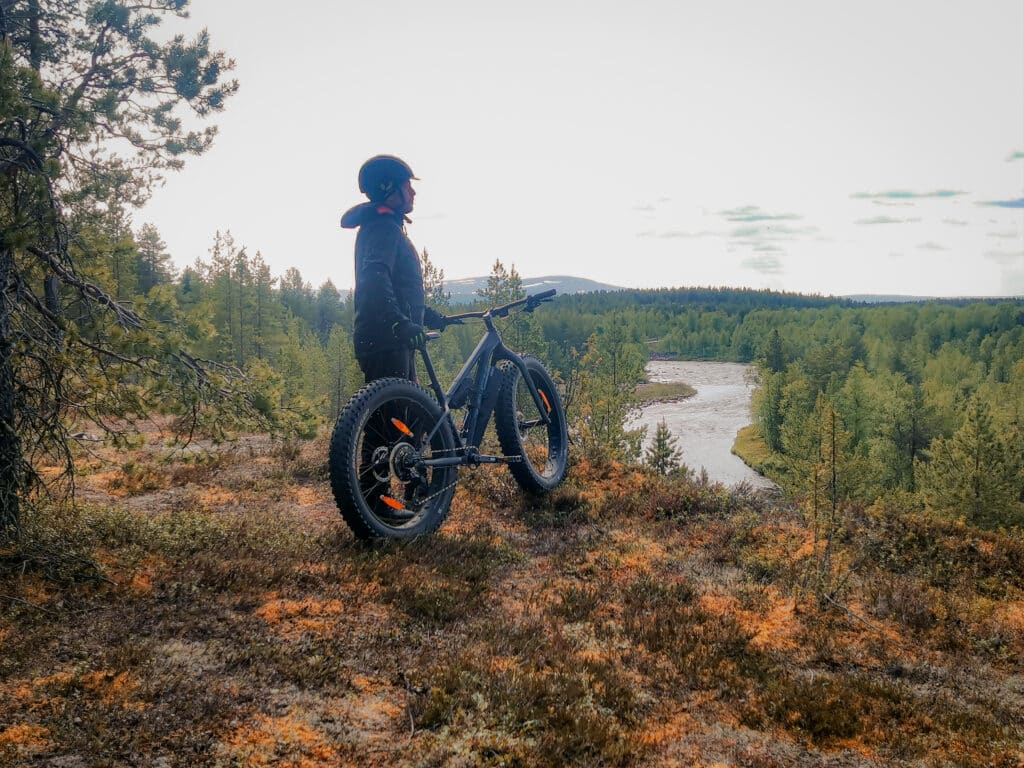 A man standing with a bike a river and fells on the backround during electric fatbike tour in Hetta Enontekiö Lapland.