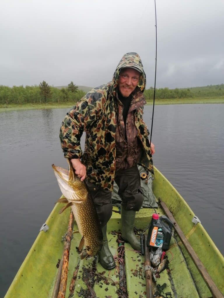 A man is standing in a boat on a lake, holding a pike he has just caught.