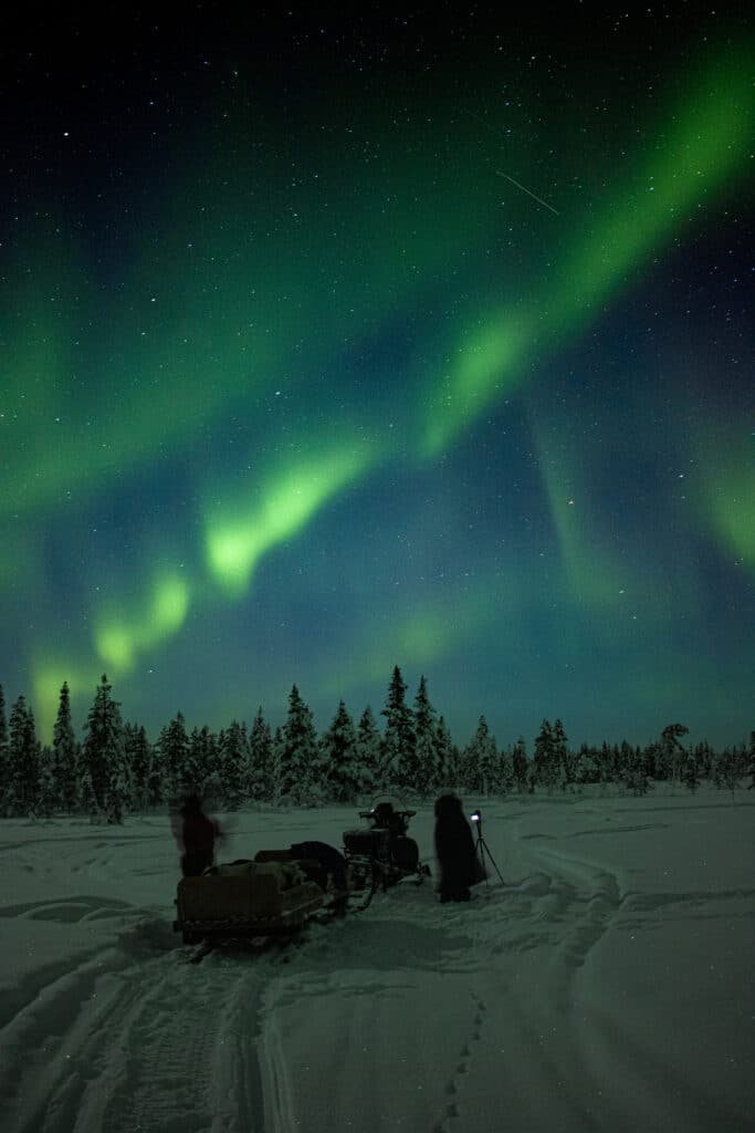 Two people standing beside a snowmobile and sleigh under the Northern Lights during the Guaranteed Northern Lights Expedition.