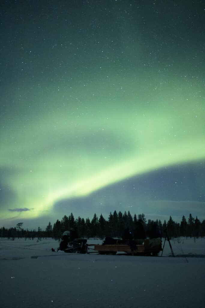 How to See the Northern Lights in Lapland by watching the aurora outdoors in the Arctic wilderness away from light pollution. People beside a wooden sleigh and snowmobile watching green Northern Lights above a snowy Lapland landscape.